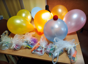 Na beżowym stole leżą różnokolorowe balony. Na jednym z nich leży czerwony nos klauna. Obok ułożone są torebki z upominkami.