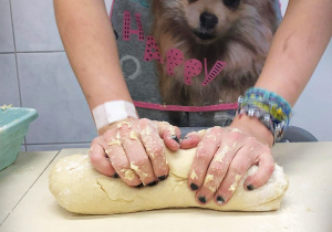 Na zdjęciu ręce dziewczyny zagniatają ciasto. Dziewczyna ma pomalowane na czarno paznokcie, na lewej ręce ma bransoletki, ubrana jest w fartuszek z brązowym pieskiem.