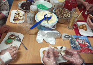Brązowy stol, na nim serek mascarpone, posypki i ozdoby cukiernicze. Przy stole siedzą dzieci i samodzielnie ozdabiają słodkie babeczki.