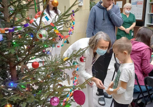 Na zdjęciu po lewej stronie stoi ubrana choinka, obok Pani w maseczce i białym ubraniu podaje chłopcu rękę. Obok stoi mężczyzna w niebieskiej bluzie, w tle brązowo żółta ściana, kobieta i dziecko siedzące przy stole.
