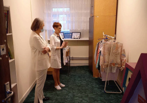 Na zdjęciu pokój pomalowany jasną farbą, po lewej stronie dwie kobiety w białych strojach. Po prawej stronie wieszak z ubraniami i brązowa szafa. W tle okno z firanką, na parapecie kolorowa praca w ramce i kartka z napisem UNICEF.
