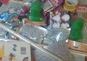 Na zdjęciu materiały plastyczne: brokatowa srebrna i złota pianka, srebrne koraliki, kleje, brokaty, folia do pakowania, zielona włóczka, sznurek, taśma klejąca, kolorowe brokaty, srebrny spray, foremki do ciastek oraz zestawy filamentów do długopisów 3d.