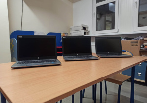 Na zdjęciu sala z jasnymi ścianami, w tle dwa okna. Z przodu brązowy stół, na nim trzy otwarte, czarne laptopy.