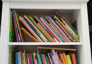 Biały regał z książkami dla dzieci. Nad regałem napis na zielonym tle "Biblioteczka dla małych i dużych pacjentów. Tutaj możesz wypożyczyć książkę, zabrać jeśli bardzo ją polubisz, przynieść swoją przeczytaną.
