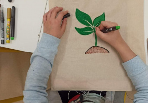 Zdjęcie przesdstawia dziecko ozdabiające płócienną, ekologiczną torbę na zakupy. Motywem pracy jest zielona roślina...