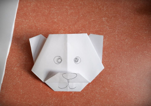 Na pomarańczowym blacie stołu leży główka misia złożona techniką origami z białej kartki papieru.