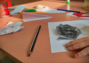 Na pomarańczowym blacie stołu widać rozsypane kredki, głowę misia zrobioną techniką origami i rękę dziecka wykonującego pracę z czarną główką misia.