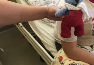 Na zdjęciu widać jak mały pacjent dostaje w prezencie misia zrobionego z włóczki, w czerwonych spodenkach i białym szaliczku.