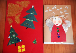 Na biurku leżą dwie kartki do św. Mikołaja. Z lewej strony na czerwonym dwie ubrane choinki, z prawej aniołek w czerwonej sukience ze srebrnymi skrzydełkami.
