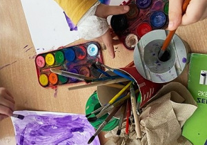 Ręce dzieci malujących farbami. Na stole leżą farby, pędzelki, woda w kubeczku i papier do wycierania pędzelków.