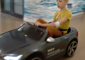 Chłopiec w szarym miniaturowym samochodzie, jedzie szpitalnym korytarzem.
