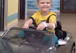 Uśmiechnięty chłopiec jedzie szarym miniaturowym samochodem, korytarzem szpitalnym.