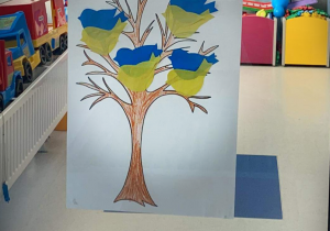 Do szyby w drzwiach przyklejona jest praca plastyczna, na której znajduje się drzewo z siedzącymi na nim czterema ptakami w kolorach niebieskim i żółtym tak jak ukraińska flaga.