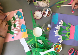Blat stołu na którym dzieci wykonują prace plastyczne przedstawiające przebiśniegi wykonane z pasków kolorowego papieru. Na stole leżą paski kolorowego papieru i klej w pojemniczku.