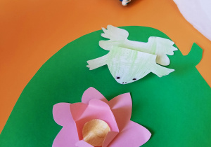 Praca plastyczna wykonana z kolorowego papieru. Na zielonym liściu jest przyklejony różowy kwiatek i żaba pokolorowana na zielono.