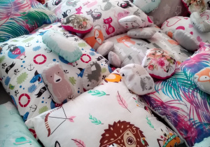 Dziecko stoi przy rozłożonych, kolorowych poduszkach w różne wzorki: jeżyki, dziewczynki, listki, miśki i różnobarwne wzory.