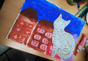 Stół, na środku praca plastyczna namalowana farbami, przedstawiająca trzy brązowe domy i szaro-złotego kota. Nad domami granatowe niebo. Obok pracy leżą kredki i klej.