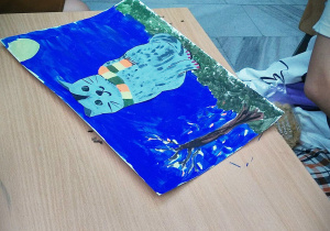 Stół, na stole praca plastyczna namalowana farbami. Na kartce namalowane granatowe niebo, brązowe drzewo, ciemnozielona trawa, na której siedzi szaro czarny kot w kolorowym szaliku. Przy stole siedzi dziecko w jasnej koszulce.