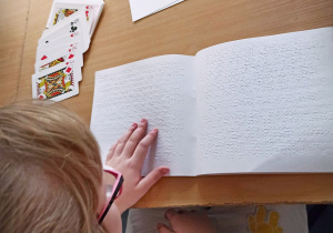Ławka, na środku leży książka napisana pismem Braille'a. Obok książki, po lewej stronie leżą karty do gry. Na dole zdjęcia jest dziewczynka w okularach, lewa ręką dotyka książki.