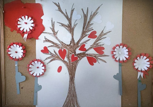 Na tablicy korkowej jest praca z narysowanym drzewem na którym zamiast liści są czerwone serduszka.Wokół pracy przyklejone są kotyliony z doczepionymi łodyżkami, tworząc razem obraz kwiatów.