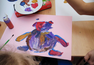 Dzieci malują farbami misie. Na pierwszym planie rysunek z wielokolorowym misiem.