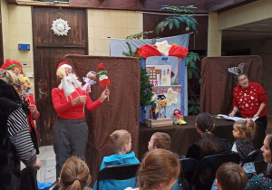 Na zdjęciu dzieci oglądające przedstawienie. W tle brązowe parawany, kolorowa scena, Panie trzymające kukiełki, po prawej stronie Pani w opasce na głowie czyta tekst przedstawienia.