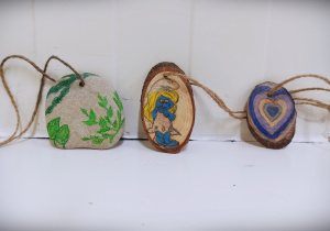 Trzy wisiorki na białym tle. Na szarym kamieniu namalowane zielone rośliny, na drewienku namalowana niebieska postać Smerfetki, na drugim drewienku fioletowo niebiesko żółte serce.