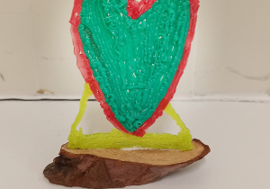Zielono czerwone serce wykonane długopisem 3 D. Serce stoi na małym drewienku.