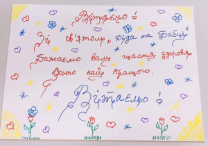 Na zdjęciu życzenia dla Babć i Dziadków w języku ukraińskim. Tekst napisany czerwonym i niebieskim długopisem na białym tle.