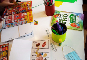 Biały stół, ręce dzieci, na środku mazaki, kleje, linijka, nożyczki i książka "Jak być eko". Na dole zdjęcia lodówka wykonana z papieru z przyklejonymi produktami wyciętymi z gazetki. Po lewej stronie dzieci wycinające produkty z gazetek reklamowych.