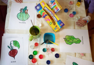 Stol, na nim farby plakatowe, ręce dzieci i palowane farbami kolorowanki przedstawiające różne warzywa i owoce .