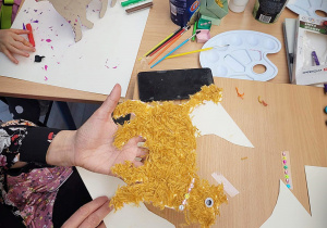 Na zdjęciu stół z różnymi materiałami plastycznymi. Na pierwszym planie jest wycięty z papieru kot, ozdobiony ruda włóczką. W tle dzieci malują koty farbami.