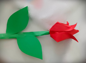 Na białym tle leży czerwony kwiatek zrobiony techniką origami z zieloną łodygą i dwoma liśćmi.