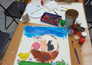 Stół, przy nim dzieci malujące i przyklejające figurki z masy solnej. Figurki przedstawiają symbole wielkanocne. Na stole leżą farby, pędzelki i naklejki.