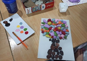 Na zdjęciu stół, pudełko z małymi kamykami, kolorowe farby i dwie prace plastyczne przedstawiające drzewa, wykonane z kamyków pomalowanych farbami.