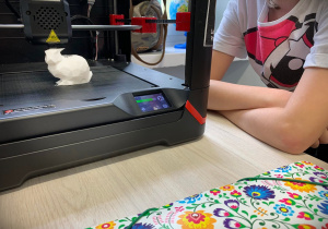 Dziecko w białej koszulce z kolorowymi wzorkami siedzi i czeka na wydrukowanie zajączka w drukarce 3D.