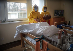 Sala z dwoma brązowymi lóżkami, na obu leżą pacjenci. Przy łóżkach dwie osoby przebrane w żółte stroje kurczaków. W rękach maja koszyczki z upominkami.
