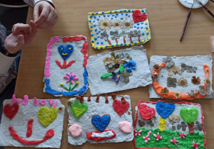 Na zdjęciu stół, na nim kolorowe prace dzieci wykonane na papierze czerpanym. Po lewej stronie ręce dzieci.
