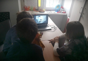 Dzieci siedzące przy zestawionych stołach. Po lewej siedzi dwóch chłopców, po prawej jeden. Przed nimi stoi laptop w którym wyświetlany jest oglądany przez chłopców film.