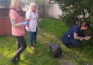 Na zielonej trawie leży czarny worek. Polewej stronie zdjęcia stoją panie, jedna w różowym fartuchu, druga w białym. Po prawej stronie zdjęcia pani w niebieskim fartuchu kuca zbierając śmieci.