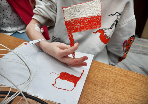 Dziecko siedzi przy beżowym stole, na którym, leży folia z wykonaną długopisem 3D flagą Polski. Taką samą flagę dziecko trzyma w ręku.