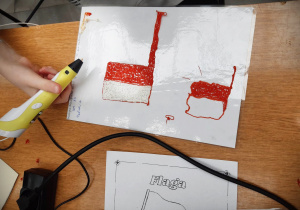 Przy beżowym stole, na białej podkładce, dziecko wykonuje żółtym długopisem 3D, dwie biało- czerwone flagi.