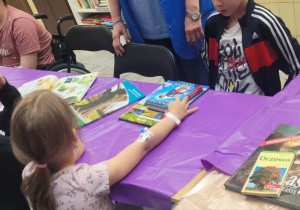 . Na zdjęciu stoły przykryte fioletową folią, przy stołach siedzą dzieci, na stole leżą książki o drzewach.