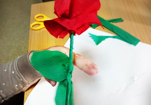 Ręka dziecka, na tle beżowego stołu, trzyma zrobioną przez siebie czerwoną różyczkę z bibuły na patyczku.
