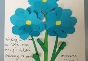 laurka na Dzień Mamy. W czerwonym wazonie wklejone są niebieskie kwiaty.Po lewej stronie napisane odręcznie zyczenia dla mamy.