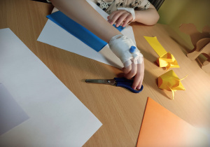 Widoczne są ręce dziecka wykonującego żółtego tulipana techniką origami. DZiecko siedzi przy jasno brązowym stole, na prawej ręce ma założony wenflon, obwiązany bandażem.
