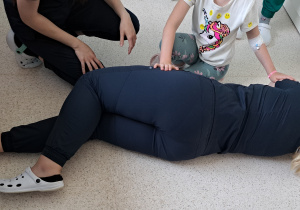 "bezpieczna pozycja boczna" - studentka w granatowym ubraniu leżąca na podłodze na boku (tyłem do nas), obok klęczy dziewczynka i studentka, która nadzorowala położenie w pozycji bezpiecznej