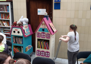 Na zdjęciu niebiesko różowy Bajkowóz z książkami, przy nim dziewczynka. Na dole zdjęcia widać głowy dzieci.