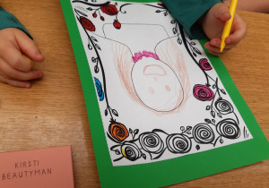 Brązowy stół, na nim ręce dziecka. Dziecko rysuje portret mamy, postać ma brązowe włosy i brązowe oczy. Na szyi namalowany różowy naszyjnik.