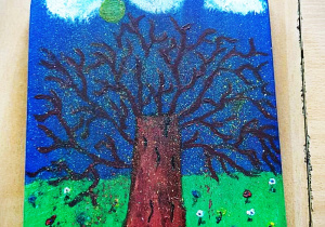 Praca plastyczna namalowana farbami na desce kuchennej. Praca przedstawia brązowe drzewo na tle zielonej trawy i niebieskiego nieba. Na trawie kolorowe kwiaty, ciemna droga, na niebie białe chmury i żółte słońce.